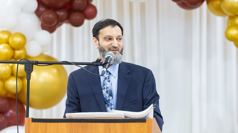 Dr. Ramzan Shahid standing behind a microphone at a podium and smiling
